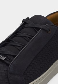 Close-up van een zwarte sneaker met geweven textuur, dikke zwarte veters en een beige zool op een lichte achtergrond.