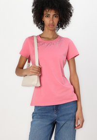 Rosa Baumwoll-T-Shirt mit kurzen Ärmeln, Rundhalsausschnitt und dekorativen Strasssteinen, die in einem Sonnenstrahlmuster um den Ausschnitt angeordnet sind, kombiniert mit blauen Jeans.
