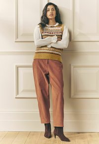 Kuldse ja mustrilise tooniga kootud vest valge pitsvarrukatega pluusi peal, kombineeritud roostepunaste sametpükstega ja pruunide pahkluu saapadega.