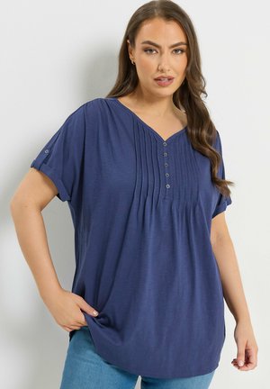 Frau trägt ein locker sitzendes, kurzärmeliges marineblaues Hemd mit Knöpfen und Falten, kombiniert mit hellblauen Jeans, steht vor einer schlichten Wand.