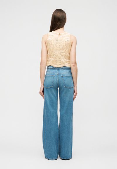 Femme aux longs cheveux bruns, debout de dos, portant un haut sans manches beige en crochet et un jean bleu taille haute à jambes larges.