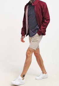 Chaqueta bomber burdeos, camisa de rayas azul marino, pantalones cortos beige, zapatillas blancas. Tatuaje en la parte inferior de la pierna. Tejido ligero, corte casual, interior en contraste.