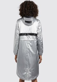 Srebrny, lekki płaszcz przeciwdeszczowy z kapturem, elastycznymi mankietami i czarną taśmą z logo na plecach. Gładka, refleksyjna tekstura. Pełnej długości.