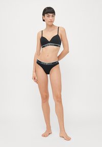 Zwart lingerie set bestaande uit een beha met verstelbare banden en een v-hals ontwerp, gecombineerd met bijpassende low-rise slipjes. Logo-accenten in het wit.