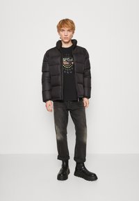 Schwarze Daunenjacke mit hohem Kragen, schwarzes T-Shirt mit grafischem Design in bunten Kreisformen, dunkle Jeans und klobige schwarze Stiefel.