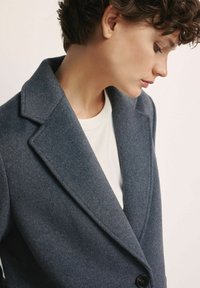 Osoba o krótkich kręconych włosach, ubrana w szary wełniany płaszcz na białą koszulę, patrząca w dół na prostym, jasnym tle.