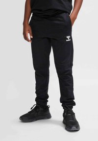 Schwarze Jogginghose mit schmaler Passform, elastischem Bund und Bündchen an den Knöcheln. Mit einem weißen Logo auf dem linken Oberschenkel. Getragen mit schwarzen Turnschuhen.