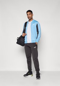 Jachetă cu fermoar de culoare albastru deschis, cu accente negre, tricou alb, pantaloni negri și pantofi sport negri. Poartă o geantă de sport neagră.