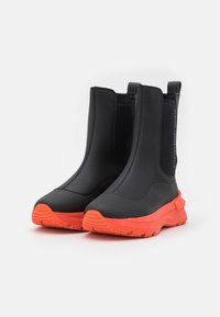 Czarne botki za kostkę z gładkiego materiału, z okrągłym noskiem, wyraźnymi pomarańczowymi podeszwami i elastycznymi bocznymi panelami, ułatwiającymi zakładanie.