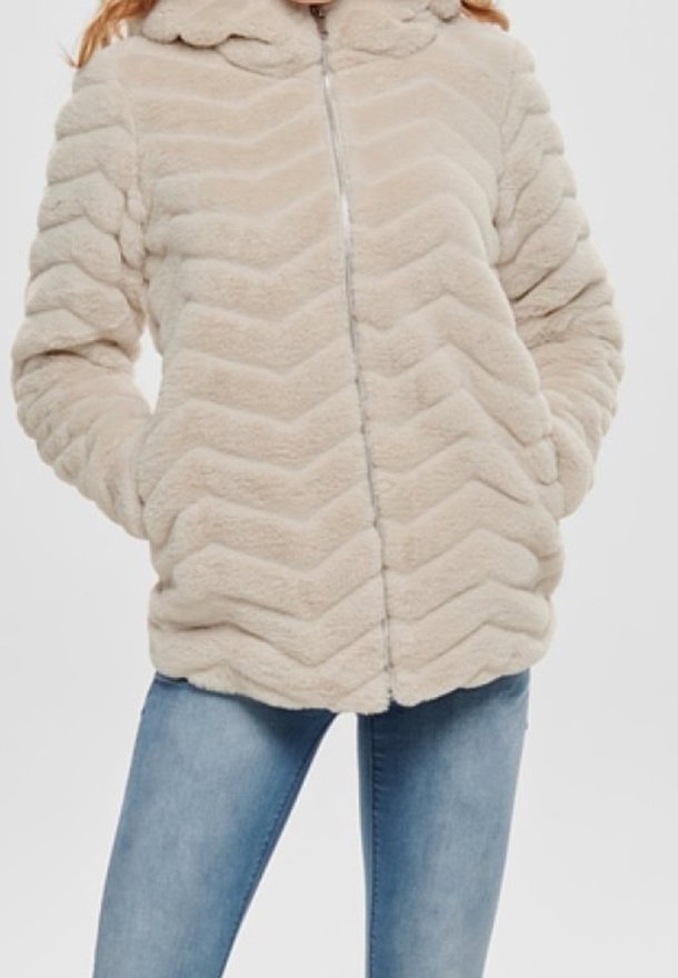 Beżowa kurtka z futerka sztucznego w wzór zygzakowy, z zamkiem z przodu i kapturem. Prezentowana z jasnoniebieskimi dżinsami. Miękki, strukturalny materiał.