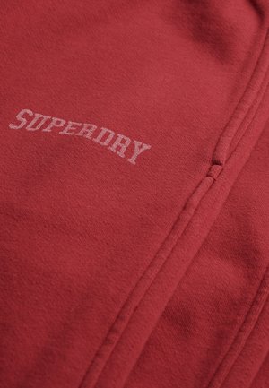 Sudadera roja de algodón, con el nombre de la marca "SUPERDRY" en relieve en gris claro. Diseño simple con acentos mínimos.