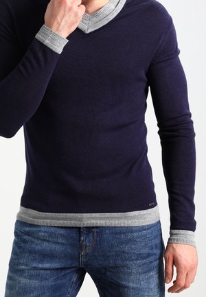 Mann trägt einen mehrschichtigen Pullover in Marineblau und Grau mit blauer Jeans, Hand nahe am Kinn in nachdenklicher Pose.