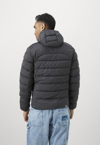 Mand iført en sort polstret hættejakke og lyseblå jeans med et logo på baglommen, der står med ryggen til kameraet.