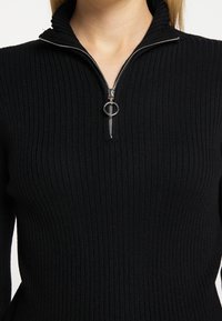 Zwarte geribbelde pullover met een hoge kraag en zilveren rits. Heeft een getailleerde pasvorm en een gestructureerde stof voor extra detail.