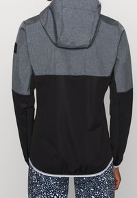 Veste grise et noire avec capuche, présentant un design épuré et un tissu texturé. Des sections contrastées mettent en valeur le haut du dos et les manches.