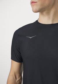 Camiseta deportiva negra hecha de una tela suave y ligera. Presenta un cuello redondo y un sutil logo reflectante en el pecho. Mangas cortas.