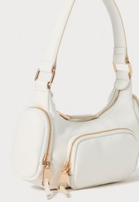 White faux leather handbag with a curved design, two zippered pockets, and gold hardware accents. Smooth texture, compact shape.