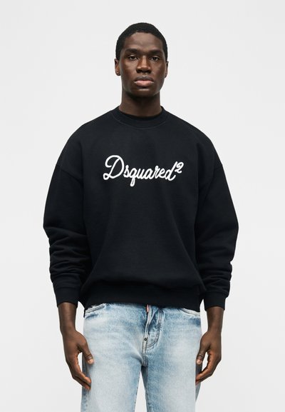 Νέος άνδρας που φοράει μαύρο φούτερ με το λογότυπο "Dsquared²" και ανοιχτόχρωμο τζιν, στέκεται μπροστά σε απλό λευκό φόντο.