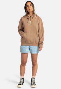 Brun hoodie med ficka framtill och snoddar, med en liten logotypdesign. I kombination med ljusa jeansshorts och svarta högskurna sneakers.