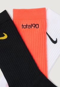 Trzy złożone ribbed skarpetki: czarne z żółwym logo Nike, neonowe pomarańczowe z napisem "total 90" oraz białe z czarnym logo Nike.