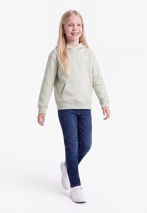 Smilende ung pige, der går, iført en lys grå hættetrøje, blå jeans og hvide sneakers på en ensfarvet hvid baggrund.