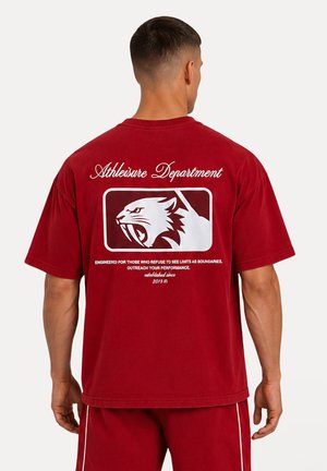 Homme portant un t-shirt rouge et un short assorti, dos montrant un logo blanc avec un tigre à dents de sabre rugissant et texte "Athleisure Department".