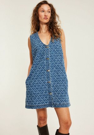 Kobieta z kręconymi włosami w niebieskiej, bezrękawnika sukience jeansowej w wzory, z przodu z guzikami i czarnych botkach do kostek, stojąca na neutralnym tle.