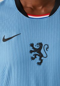 Heleda sinine spordisärk, millel on tekstuurset kangast, must Nike logo ja must lõvi embleem alumises ees. Omab musta kaelust.