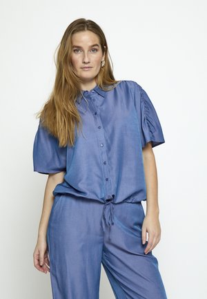 Žena s dugom smeđom kosom, odjevena u široku plavu košulju s dugmadima i skupljenim kratkim rukavima te hlače u istom stilu, stoji ispred bijele pozadine.