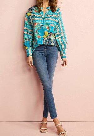 Lyseblå blomstret bluse med v-hals og lange ærmer, kombineret med tætsiddende lyse denimjeans og hvide sandaler med stropper.