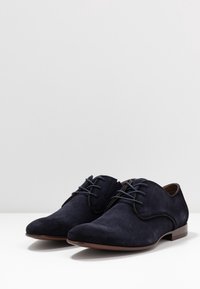 Chaussures habillées en daim bleu marine avec un bout rond, des lacets plats et une semelle en cuir brun lisse. Design minimaliste sans ornement.
