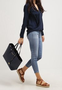 Marineblauwe draagtas met dubbele handgrepen, gecombineerd met een donkerblouse, lichtblauwe spijkerbroek en bruine sandaal met sleehak en bandjes.