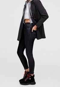 Jean skinny noir taille haute, associé à un haut blanc court et un sweat à capuche gris, complété par des baskets noires avec des accents rouges.