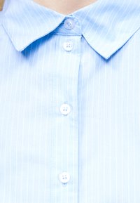 Lichtblauw overhemd met dunne witte verticale strepen, een puntkraag en vier witte knopen aan de voorkant dichtgeknoopt.