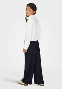 Camicia bianca corta con maniche lunghe, abbinata a pantaloni larghi blu navy e sneakers beige. Tessuto liscio, vestibilità su misura e colletto arrotondato.