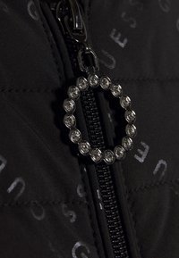 Zbliżenie na czarny zamek błyskawiczny z okrągłym uchwytem ozdobionym przezroczystymi rhinestonami na czarnej tkaninie z subtelnie nadrukowanymi literami.