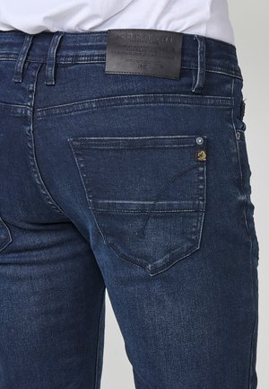 Vue arrière d'un jean en denim bleu foncé porté avec une chemise blanche, comportant une étiquette de marque en cuir noir et une poche arrière avec des détails de couture.