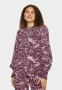 Donna con capelli ricci che indossa una blusa e pantaloni a maniche lunghe a pois maroon e rosa coordinati, in piedi contro uno sfondo bianco semplice.