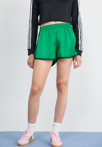 Green athletic shorts with black trim and an Adidas logo, paired with a black cropped long-sleeve shirt and pink sneakers.