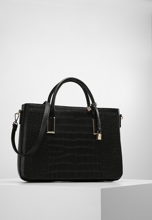 Sac à main en cuir noir à texture crocodile, deux poignées, bandoulière amovible et petit détail de cadenas doré sur une plateforme blanche.
