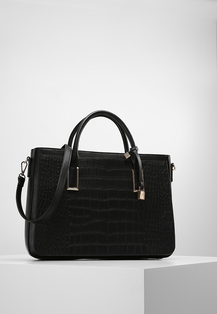 Sac à main en cuir noir à texture crocodile, deux poignées, bandoulière amovible et petit détail de cadenas doré sur une plateforme blanche.