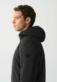 Schwarze Pufferjacke mit Kapuze, glatter Textur und einem runden Logo auf der linken Schulter. Die Seitenansicht hebt das schlanke Design hervor.