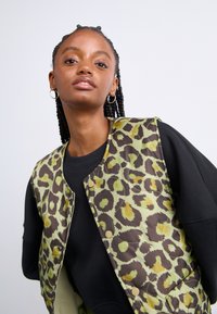 Polstret vest med et grønt og brunt leopardmønster. Den har trykknapper og er lagdelt over en svart langermet topp.