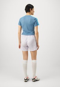 T-shirt de sport bleu clair à manches courtes, associé à un short blanc. Le mannequin porte des chaussettes blanches jusqu'aux genoux et des crampons noirs avec des accents rouges.