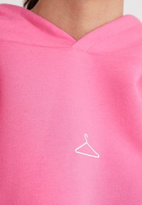 Sudadera con capucha rosa hecha de tela suave, con escote en v y un pequeño gráfico blanco de un colgador en la parte delantera. Textura suave y diseño sencillo.