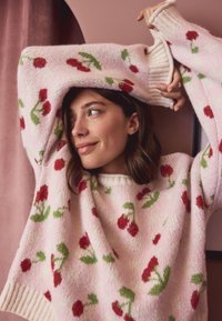 Maglione rosa lavorato a maglia con motivi di fiori rossi e foglie verdi. Polsini e scollo a coste, tessuto morbido con vestibilità leggermente ampia.