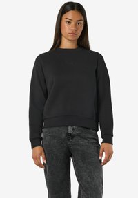 Mujer joven con el cabello oscuro y liso, viste un sudadera negra sencilla y unos jeans negros desgastados, de pie contra un fondo blanco.