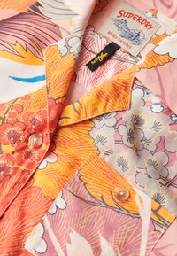 Chemise à manches courtes avec des motifs floraux en rose, orange et jaune. Elle présente un col pointu, des boutons et un tissu en coton doux.