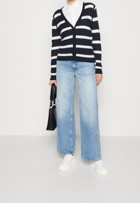 Cardigan a righe blu navy e bianche, design a V, indossato sopra una camicia bianca con colletto. Jeans a gamba larga blu chiaro e sneakers bianche. Borsa nera.