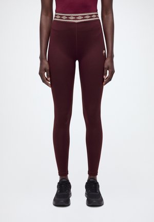 Un modèle porte un legging bordeaux pleine longueur à taille imprimée et des baskets noires, et se tient devant un fond uni.
